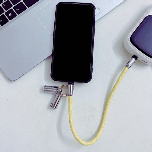 Smartphone with a yellow phone wrist strap that is also a charging cable attached, placed on a white surface with a laptop in the background.