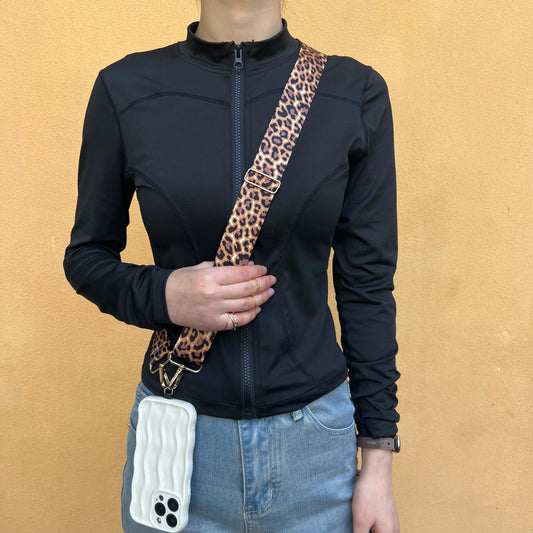 Person wearing an animal print cross body phone strap with a white cell phone case hanging from the strap. The person is wearing a black jacket and standing in front of a yellow wall. 